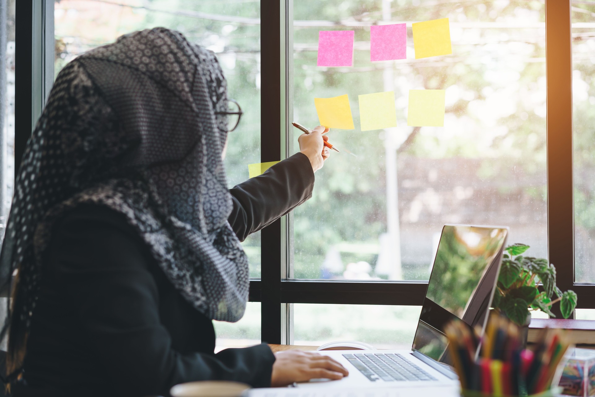 Asian muslim business woman reaching hand picking sticky notes on glass wall.